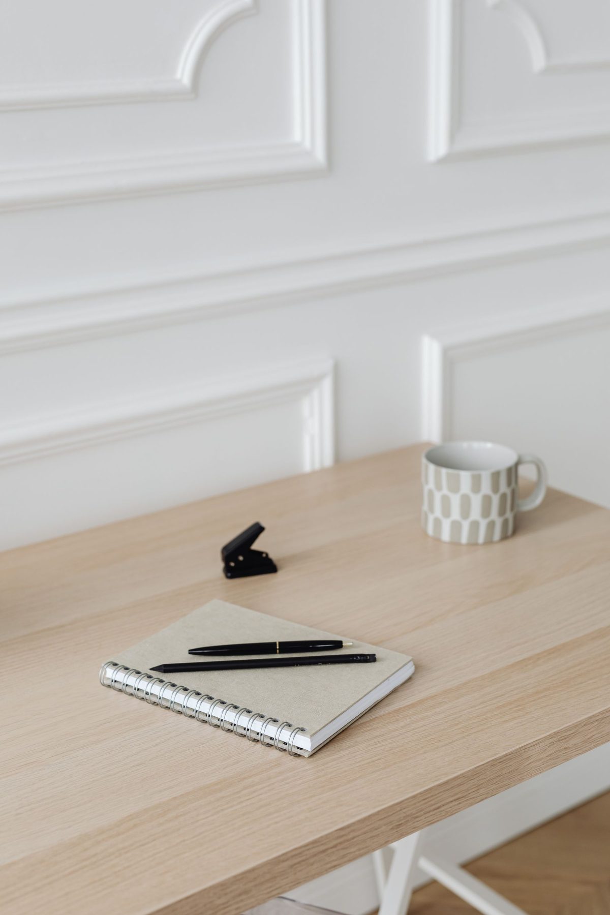 A notebook, pens and a mug on a light wooden desk